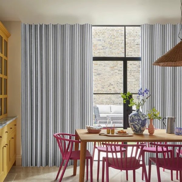 A brightly lit dining room featuring striped floor-to-ceiling curtains, a wooden table with pink chairs, and a large window looking out onto a patio.