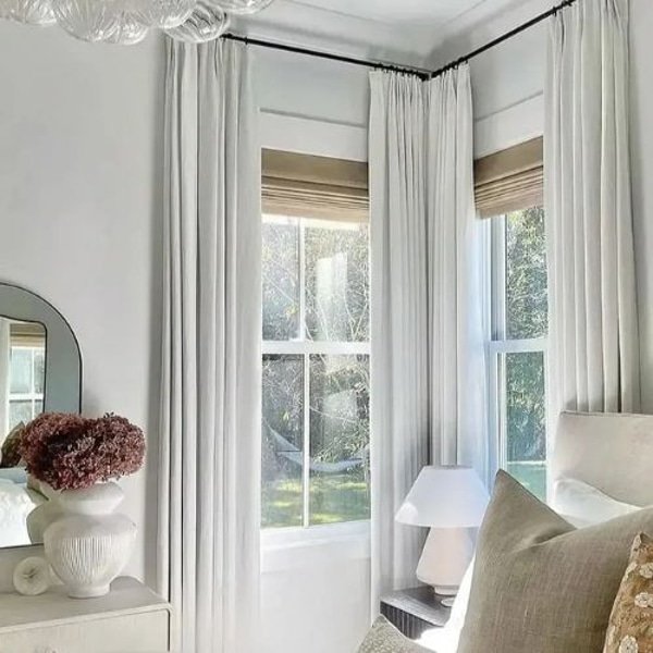 A corner of a room showing white pleated curtains hanging on a black rod over windows with tan Roman shades.