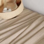 A high-angle shot of folded beige linen fabric next to a woven basket containing a glass carafe and a cotton boll.