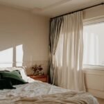 A sunlit, cozy bedroom featuring cream-colored curtains, a rumpled white bedspread, and dark green velvet pillows.