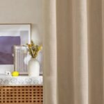 Close-up of a beige linen curtain hanging next to a white vase with yellow flowers and framed abstract art.